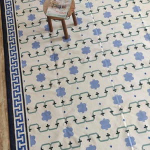 Op de afbeelding: Een groot, rechthoekig tapijt met een herhalend patroon van blauwe bloemvormen en verbindingslijnen op een crèmekleurige achtergrond. De rand is marineblauw met een Grieks sleutelontwerp. Een kleine houten kruk met een tijdschrift staat in het midden.