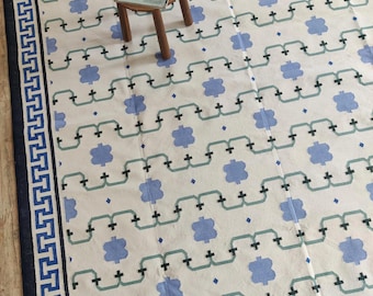 May include: A large, rectangular rug with a repeating pattern of blue floral shapes and connecting lines on a cream background. The border is navy blue with a Greek key design. A small wooden stool with a magazine on top is in the center.