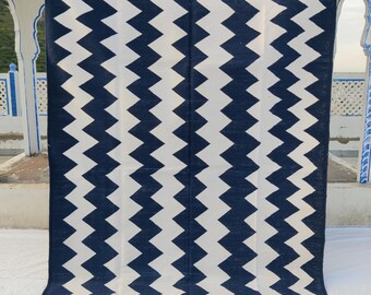 May include: A large, rectangular rug with a bold zigzag pattern. The rug features alternating bands of navy blue and white, creating a striking visual contrast. The edges have a fringe detail. This rug is suitable for home decor.
