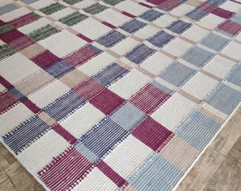 May include: A large, rectangular area rug with a geometric plaid pattern. The rug features squares in shades of white, blue, purple, green, and tan. The rug is made of a woven material and is placed on a wooden floor.