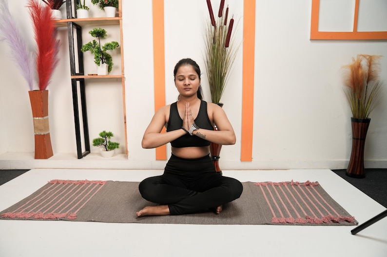 May include: A person in a black sports bra and leggings sits in a yoga pose on a grey mat with pink stripes. The background includes decorative plants, a wooden shelf, and tall vases with decorative plants.