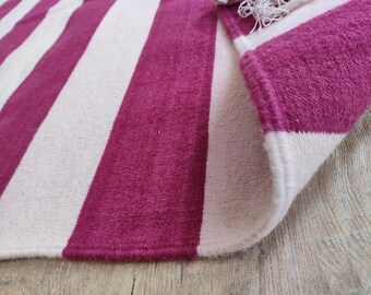 Puede incluir: Primer plano de una alfombra con anchas rayas magenta y blanco roto. El borde de la alfombra es visible, mostrando el grosor y la textura del material. La alfombra está doblada, revelando la parte inferior.