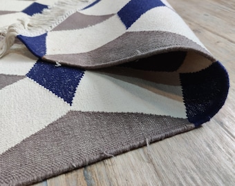 May include: A geometric patterned rug with a rolled edge. The rug features a design of blue, grey, and white diamond shapes. The rug has a fringe on one end and is made of a woven material.