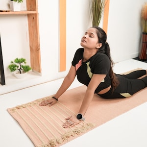 May include: A person in a yoga pose on a light pink rectangular yoga mat with a fringed edge. The person is wearing a black athletic top and leggings. A small potted plant and decorative items are in the background.