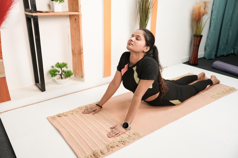 May include: A person in a yoga pose on a light pink rectangular yoga mat with a fringed edge. The person is wearing a black athletic top and leggings. A small potted plant and decorative items are in the background.