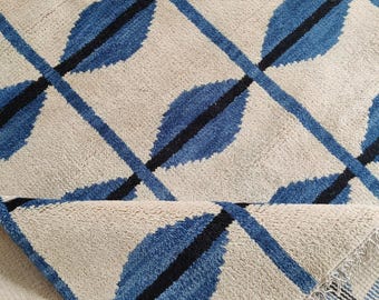 May include: A rolled-up rug with a repeating pattern of blue leaf shapes and black lines on a cream background. The rug's design features a geometric arrangement of the leaf motifs, creating a textured surface. The rug's edge is visible, showing the fringe.