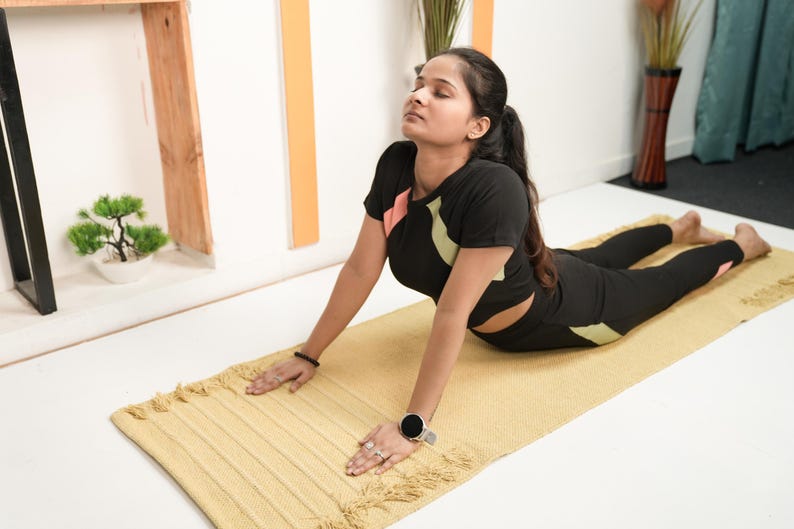 May include: A person in a yoga pose on a yellow mat. They are wearing a black athletic top with pink and green accents and black leggings. The mat is rectangular with a fringed edge. A small bonsai tree is in the background.