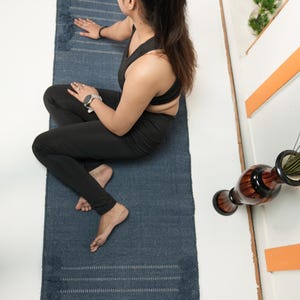 May include: A blue yoga mat with fringing. The mat has a textured surface and horizontal stripes. The mat is rectangular and appears to be made of woven material. The mat is on a white surface.