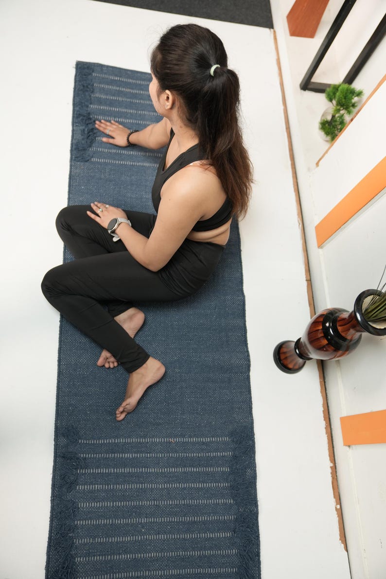 May include: A blue yoga mat with fringing. The mat has a textured surface and horizontal stripes. The mat is rectangular and appears to be made of woven material. The mat is on a white surface.