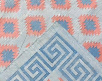 Puede incluir: Un textil de color claro con un patrón repetitivo de formas geométricas azules y coral. El borde presenta un diseño de llave griega azul. La tela está doblada, revelando el patrón y el borde.