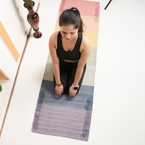 May include: A woman kneeling on a multi-coloured yoga mat. The mat has a gradient of colours, including pink, yellow, green, blue, and purple. The woman wears a black sports bra and leggings. A watch is on her wrist.