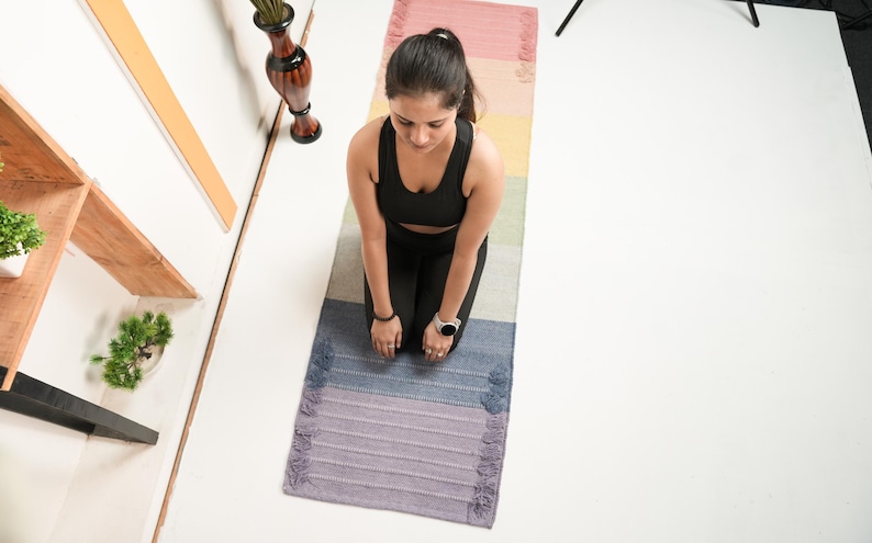 May include: A woman kneeling on a multi-coloured yoga mat. The mat has a gradient of colours, including pink, yellow, green, blue, and purple. The woman wears a black sports bra and leggings. A watch is on her wrist.