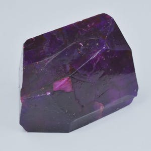 Op de afbeelding: Een gefacetteerde, kristallijne structuur in tinten diep paars en magenta. De edelsteen heeft een ruwe, natuurlijke textuur met zichtbare interne patronen en kleurvariaties. De algemene vorm is onregelmatig, met meerdere vlakke oppervlakken en scherpe randen.