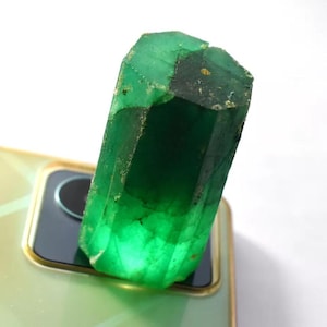 Può includere: Un cristallo di smeraldo verde vibrante, con una forma rettangolare sfaccettata. La gemma ha una texture naturale e ruvida e diverse sfumature di verde, con alcune aree traslucide. Il cristallo è posto su uno sfondo chiaro.
