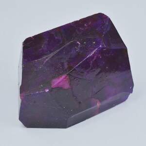 Op de afbeelding: Een gefacetteerde, kristallijne structuur in tinten diep paars en magenta. De edelsteen heeft een ruwe, natuurlijke textuur met zichtbare interne patronen en kleurvariaties. De algemene vorm is onregelmatig, met meerdere vlakke oppervlakken en scherpe randen.