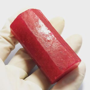 May include: A faceted, rectangular, deep red gemstone is held in a gloved hand. The gem has a polished, slightly textured surface and a geometric shape. The color is a vibrant, saturated red.