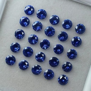 May include: A collection of twenty-one round, faceted blue gemstones arranged in a grid pattern. Each stone features a brilliant cut with multiple reflective surfaces that create a deep, vibrant royal blue color throughout the circular shape.