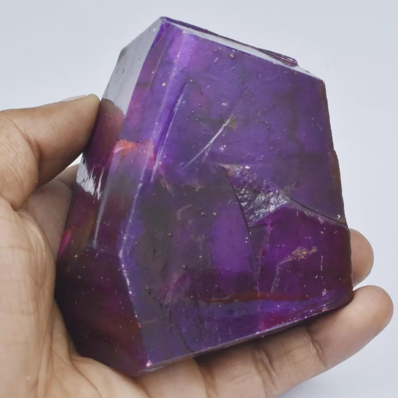 Op de afbeelding: Een gefacetteerd, dieppaars kristal in een hand. Het kristal heeft een rechthoekige vorm met meerdere schuine oppervlakken, die licht reflecteren. De kleur varieert van donker, bijna zwart, tot levendig paars.