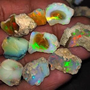 May include: A collection of raw, unpolished opal gemstones featuring irregular shapes and rough, earthy matrix exteriors. The stones display vibrant flashes of iridescent color, including bright green, orange, and blue, set against milky white and translucent base tones.