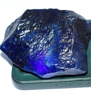 Puede incluir: Una piedra preciosa en bruto de color azul profundo con una textura cristalina irregular y bordes translúcidos. La piedra muestra tonos variados de azul oscuro y real, con una superficie rugosa sin pulir y facetas naturales.