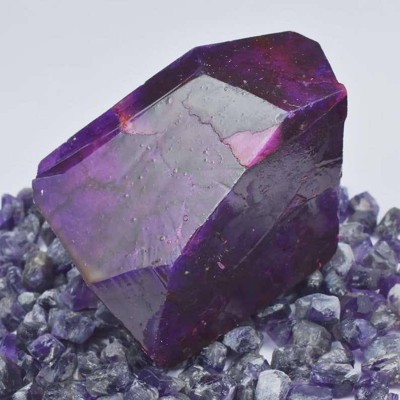 Op de afbeelding: Een grote, gefacetteerde paarse kristal met een diepe, rijke kleur, rustend op een bed van kleinere, vergelijkbare kristallen. Het hoofdkristal heeft een gepolijst, reflecterend oppervlak, dat de geometrische structuur en interne diepte laat zien.