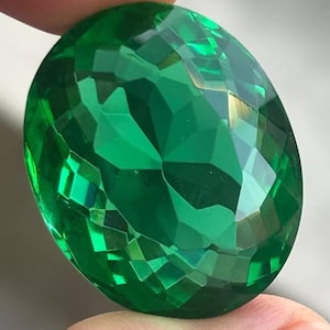 May include: A large, oval-shaped, faceted gemstone with a vibrant, translucent emerald green color. The stone features numerous geometric cuts across its surface, creating a reflective, sparkling appearance with varying shades of green light.