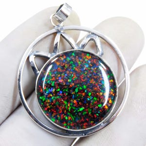 May include: A silver pendant featuring a large, round, black opal stone with vibrant flecks of red, green, and blue. The opal is set within a circular frame, with a decorative star-like design above. The pendant is held by a gloved hand.