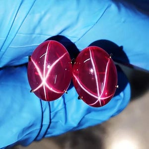 May include: Two oval-shaped, deep red star rubies, each featuring a distinct six-rayed white star pattern across the smooth, polished cabochon surface. These gemstones are set in metal prongs, displaying a vibrant, translucent crimson hue.