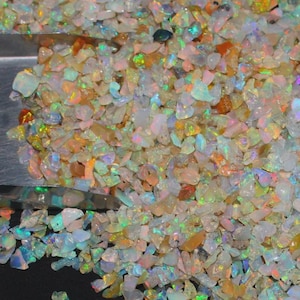Könnte beinhalten: Eine Ansammlung kleiner, unregelmäßiger Opal-Splitter mit einer durchscheinenden Basis und leuchtenden Blitzen in Grün, Orange, Blau und Pink. Eine Metallschaufel steckt teilweise in dem Haufen der Edelsteine, die auf einer dunklen Fläche liegen.