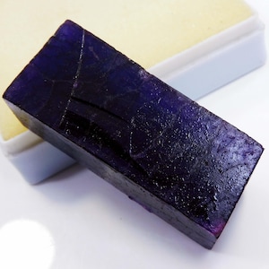 Può includere: Una gemma rettangolare, viola scuro, con una superficie ruvida e strutturata. La pietra è di un colore ricco e scuro con crepe e imperfezioni visibili. È parzialmente appoggiata su una superficie bianca e gialla.