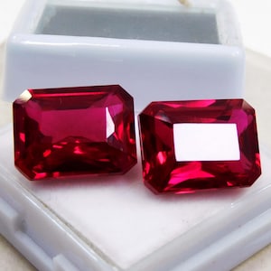 Puede incluir: Dos gemas de color rojo intenso con talla radiante. Las gemas se exhiben en una caja de plástico blanco, mostrando su color vibrante y facetas. Las piedras tienen forma rectangular con esquinas cortadas, reflejando la luz.