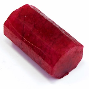 Puede incluir: Una gema de rubí en bruto y sin cortar, de color rojo intenso. La piedra de forma rectangular tiene una superficie facetada y una textura ligeramente rugosa. El rubí se muestra sobre un fondo blanco, resaltando su color y forma.