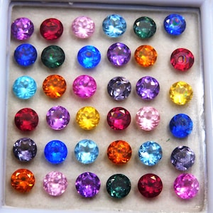 Puede incluir: Una colección de gemas redondas en varios colores, incluyendo púrpura, rojo, rosa, azul, verde, naranja y amarillo. Las gemas están dispuestas en un patrón de cuadrícula dentro de una bandeja blanca, mostrando sus vibrantes tonos y superficies reflectantes.