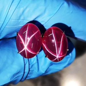 Può includere: Due gemme ovali rosso intenso con un motivo a stella di linee bianche. Le gemme sono incastonate in griffe di metallo color oro. Lo sfondo è una superficie azzurra.