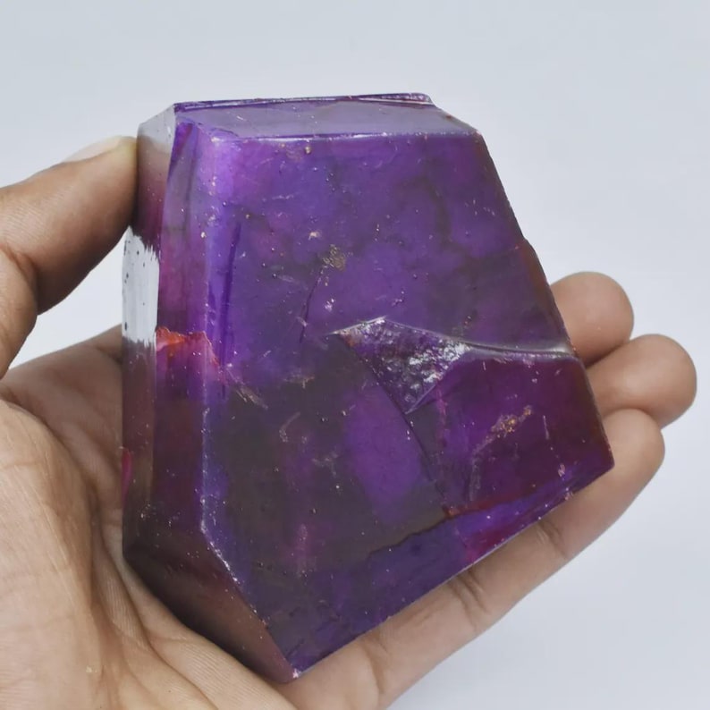 Op de afbeelding: Een gefacetteerd, dieppaars kristal in een hand. Het kristal heeft een gepolijst oppervlak met kleur- en textuurvariaties. De vorm is geometrisch, met vlakke vlakken en scherpe hoeken. Het lijkt op een edelsteen of een decoratief object.