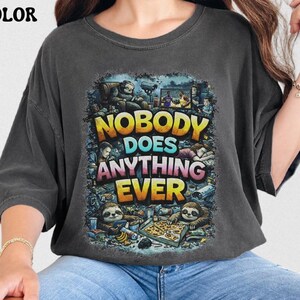 Puede incluir: Camiseta de gran tamaño color pimienta con el texto "NOBODY DOES ANYTHING EVER" en amarillo, azul y rosa. El gráfico presenta perezosos y otros personajes de dibujos animados. La camiseta se lleva con vaqueros azules.