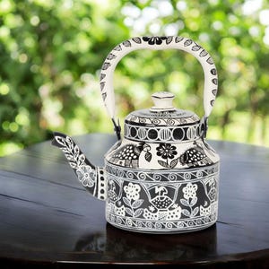 May include: Black and white hand-painted teapot with a floral and bird design. The teapot has a curved spout, a rounded body, and a white handle. The background is a blurred green.