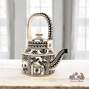 May include: Cream-colored ceramic teapot with black decorative patterns. The teapot features a curved handle, a spout, and a lid. The design includes floral and bird motifs, with the text "RajasthanRoots" in the lower right corner.