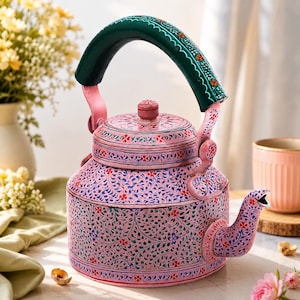 Pode incluir: Uma chaleira de metal rosa com um intrincado padrão floral pintado à mão em azul, verde e vermelho. A chaleira tem uma alça arqueada verde-escura com detalhes florais laranja, uma tampa a condizer com um puxador e um bico curvo.
