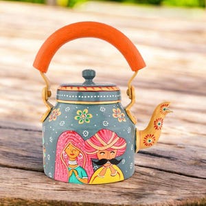 May include: Hand-painted teal teapot with an orange handle and spout. The teapot features a painted design of a couple in traditional attire, with floral accents. The handle and spout are painted with floral patterns. The teapot is sitting on a wooden surface.
