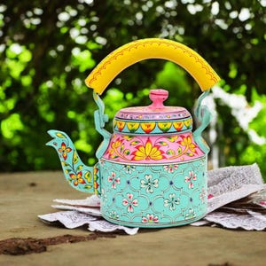 May include: A hand-painted, colorful teapot with a yellow handle and spout. The teapot features a floral design in shades of pink, blue, yellow, and green. The teapot is sitting on a wooden surface.