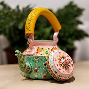 May include: A decorative teapot with a yellow handle and a lid. The teapot is painted with floral patterns in pink, green, and orange. The lid is off to the side, revealing the inside of the teapot.