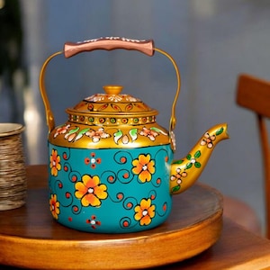 Puede incluir: Una tetera pintada a mano en color turquesa y dorado con un diseño floral. La tetera presenta flores naranjas y amarillas, un asa de color cobre y un pico dorado. La tapa y el borde también son dorados, creando una pieza vibrante y decorativa.