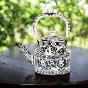 May include: Black and white hand-painted teapot with a floral and bird design. The teapot has a curved spout, a rounded body, and a white handle. The lid is white with a small knob. The teapot is sitting on a dark wooden table.