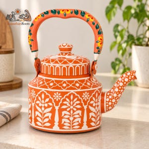 May include: An orange metal teapot featuring white hand-painted floral and geometric patterns. The handle is decorated with yellow and green floral motifs. The spout is covered in white polka dots. The text RajasthanRoots is visible.