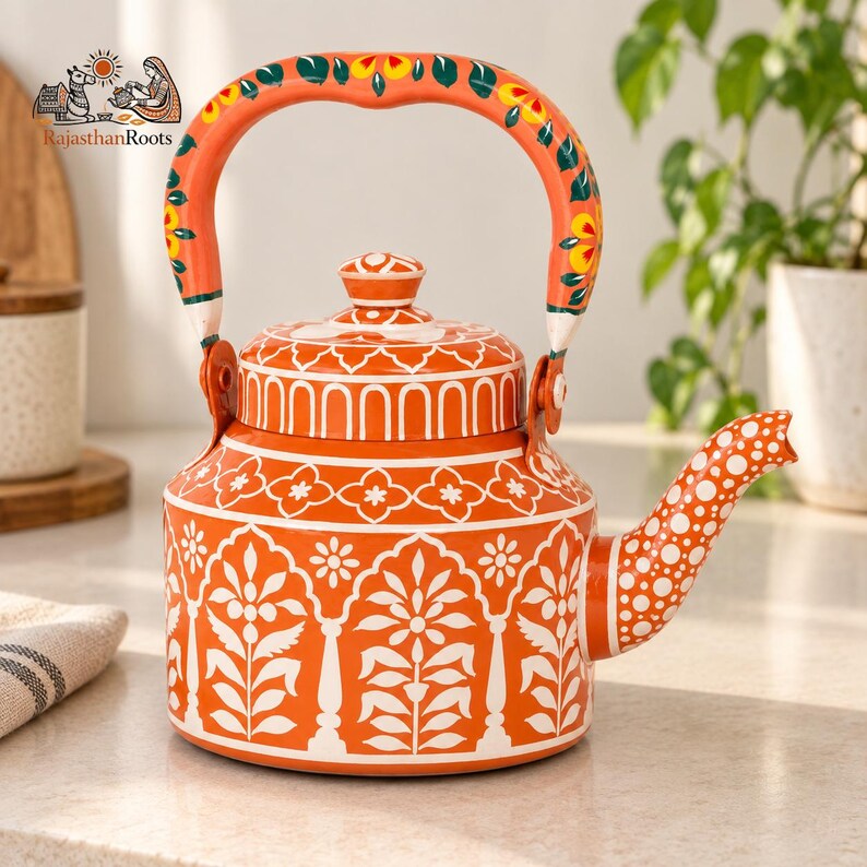 May include: An orange metal teapot featuring white hand-painted floral and geometric patterns. The handle is decorated with yellow and green floral motifs. The spout is covered in white polka dots. The text RajasthanRoots is visible.