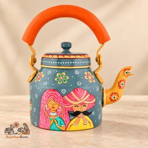 Puede incluir: Una tetera de metal color verde azulado con un diseño pintado a mano de una persona con velo rosa y otra con turbante amarillo y bigote. La tetera tiene un asa naranja, un pico amarillo con motivos florales y detalles de puntos blancos.