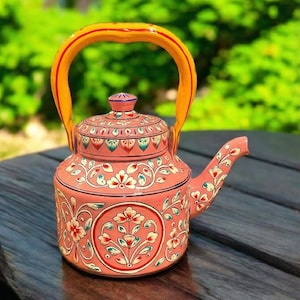 May include: A hand-painted, coral-colored teapot with a yellow handle and lid. The teapot is adorned with floral patterns in white, red, and green. The design is symmetrical, with a circular floral motif on the front and back. The teapot is on a wooden surface.