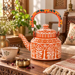 May include: A vibrant orange metal teapot featuring intricate white floral and geometric patterns across its body, lid, and arched handle. The spout is decorated with white polka dots, and the lid is topped with a small, matching orange knob.