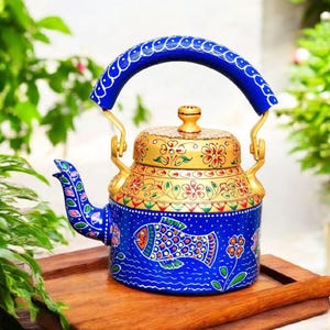 May include: A hand-painted blue and gold metal teapot with a fish design. The teapot features a curved blue handle and a lid with floral patterns. It sits on a wooden tray, with green foliage in the background.